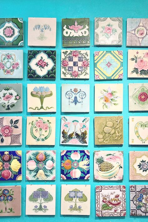 a vibrant turquoise wall is adorned with a meticulously arranged collection of vintage tiles.  each square tile showcases a unique and intricate design, predominantly featuring floral motifs, fruits (peaches, pineapples), and geometric patterns. the tiles vary in color palettes but share a consistent aesthetic, suggesting a cohesive collection likely from a similar era or style. the overall impression is one of elegant and detailed craftsmanship, showcasing a beautiful display of antique tile artistry.