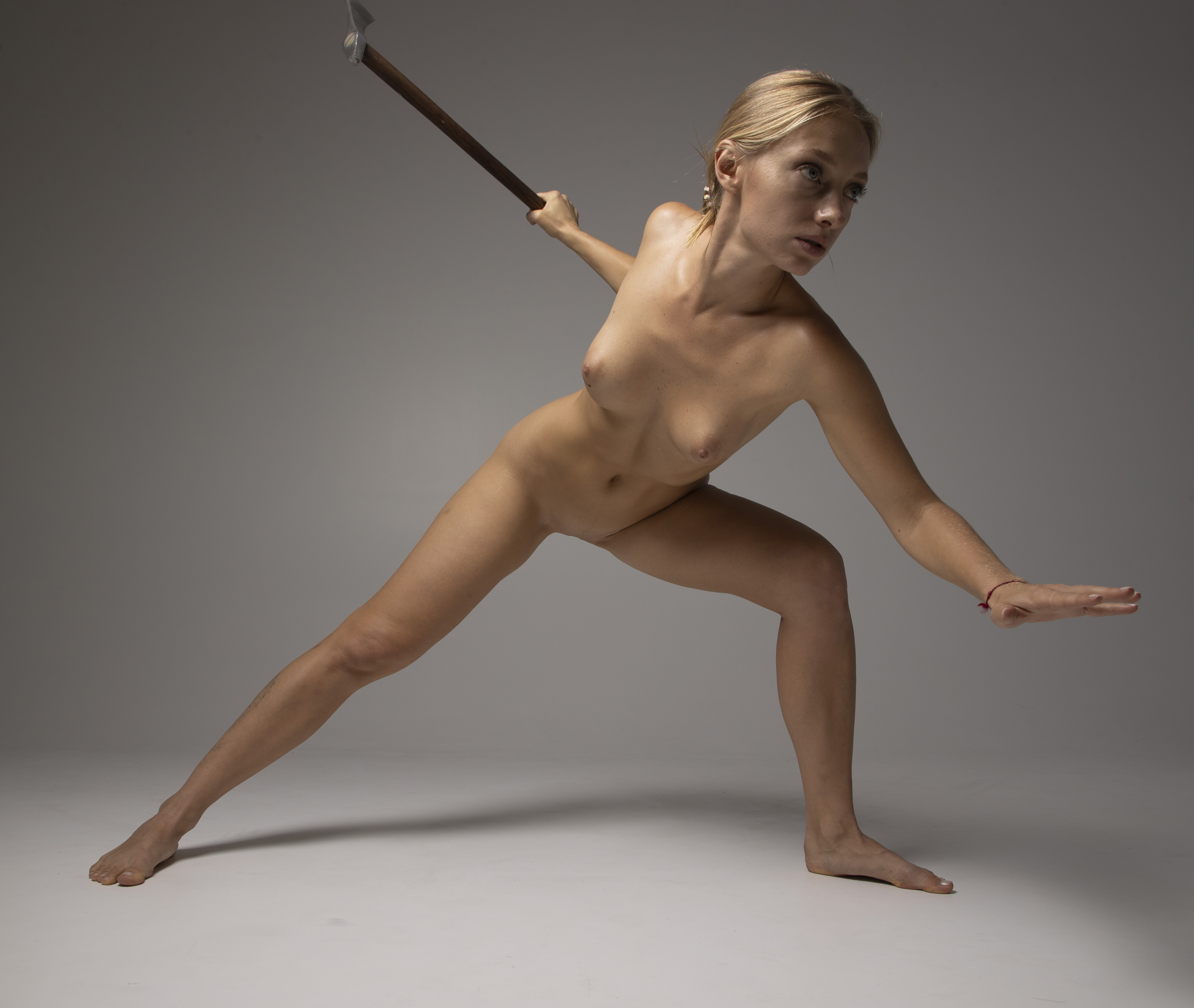 a nude woman with blonde hair is holding a spear in a studio shot. she is posed in a warrior stance, with one leg forward and the other back.