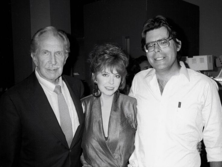 a black and white photo of three people, two men and a woman. the two men are wearing suits, the woman is wearing a dress. the man on the left has his hand on the woman's shoulder. the man on the right has his arm around the woman's waist. all three are smiling. the man on the right is wearing glasses. they are all posing for a photo.