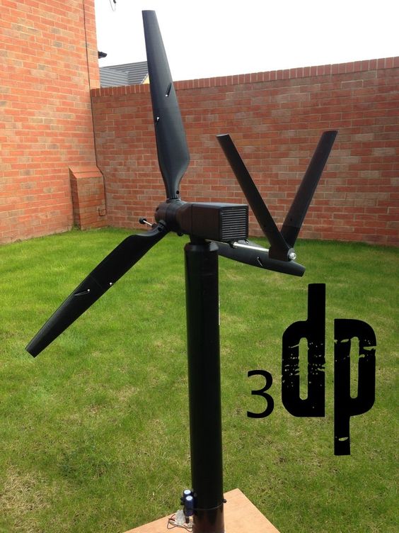 a black wind turbine with three blades is mounted on a tall black pole in a backyard. the turbine is facing the camera and is positioned in front of a brick wall. the grass is green and lush. the turbine has a small generator attached to the center hub.