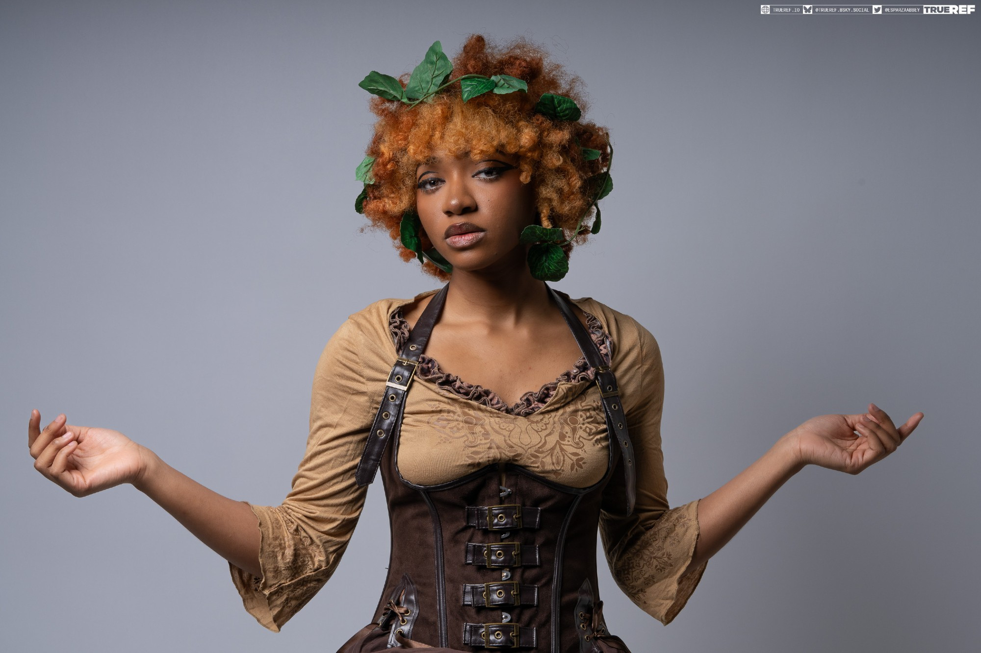 a closeup studio portrait of a young black woman with vibrant orange curly hair adorned with green leaves. she is dressed in a steampunkinspired brown corset and tan blouse.  her arms are outstretched, as if presenting something or making a gesture. her expression is serious and confident. the background is a neutral grey.