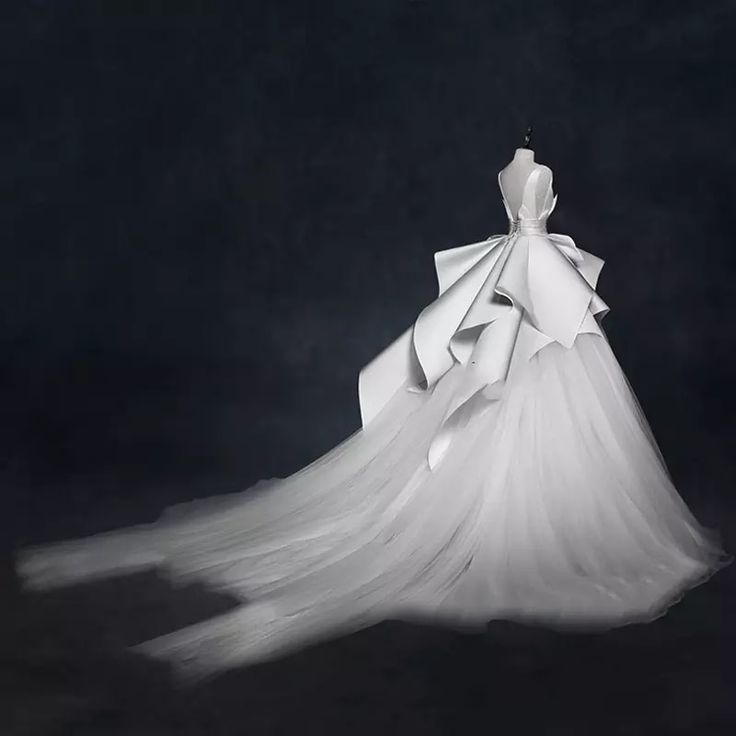 a white wedding dress with a long tulle train, a satin bodice, and large bow at the back. the dress is on a mannequin on a black background.  the dress has a lot of ruffles on the back and the train, giving it a dramatic look.