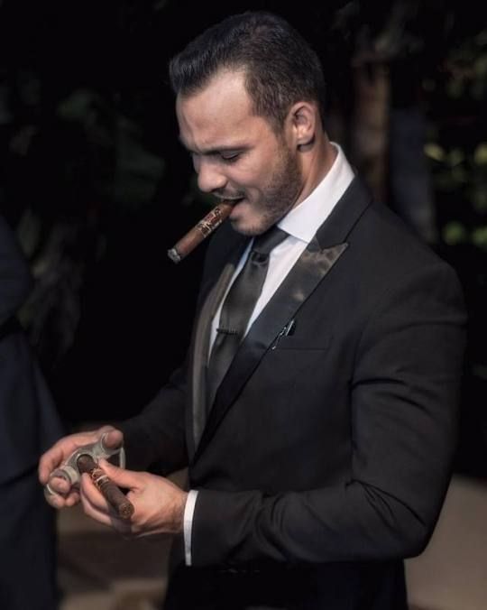 a portrait of a man wearing a black suit, tie, and tuxedo smiling while smoking a cigar.