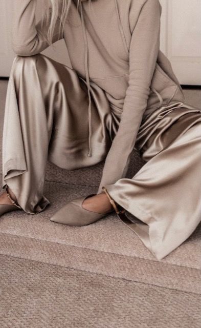 a woman is sitting on a carpeted floor. she is wearing beige satin pants, a beige hoodie and beige shoes. she is leaning forward, her left leg crossed over her right leg. her right hand is resting on her right leg.