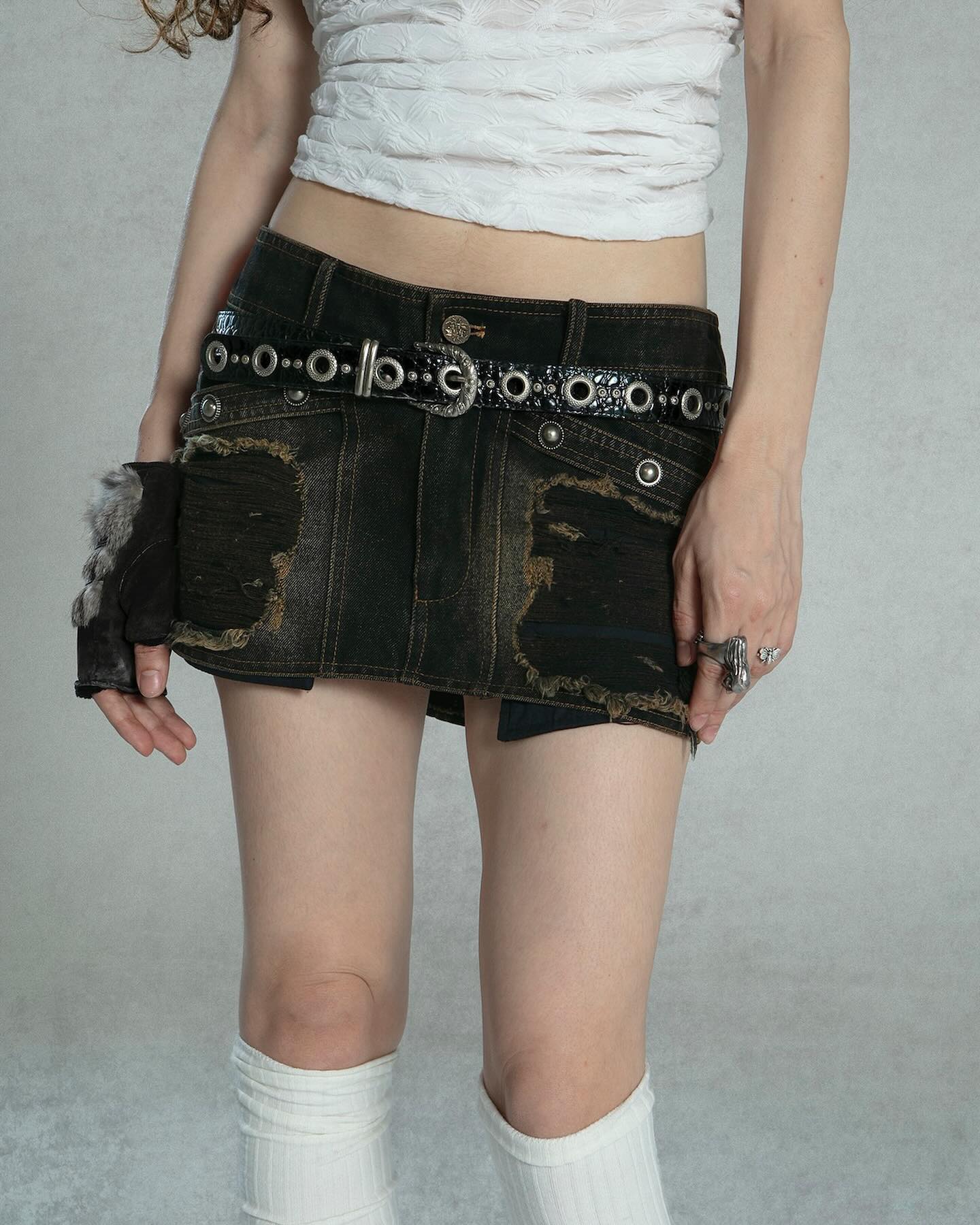 the image shows a young woman modeling a darkwash denim mini skirt with distressed detailing and visible rips.  the skirt is paired with a white cropped shirt and a black studded belt with metal eyelets. she's wearing white kneehigh socks and has some jewelry on her hands.  the setting appears to be a studio with a neutral gray backdrop. the overall style is grunge or punkinspired. the focus is on the clothing, particularly the distressed denim skirt.