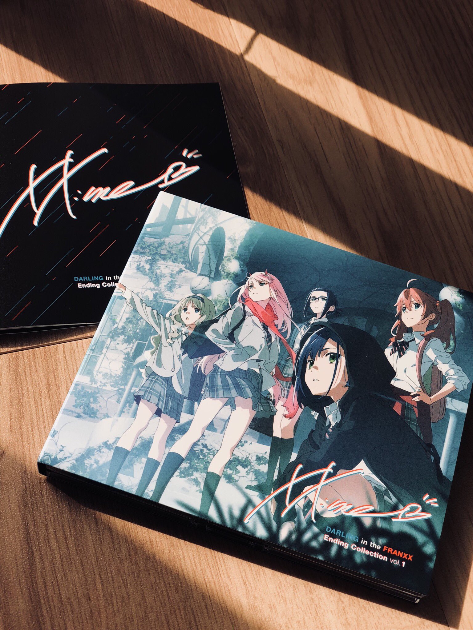 two darling in the franxx anime cds lie on a hardwood floor. the cds are part of the "ending collection," and the covers feature illustrations of the female characters in school uniforms. the cover of volume 1 is angled towards the camera, while the cover of volume 2 is partially obscured. natural light from the right casts shadows on the floor.