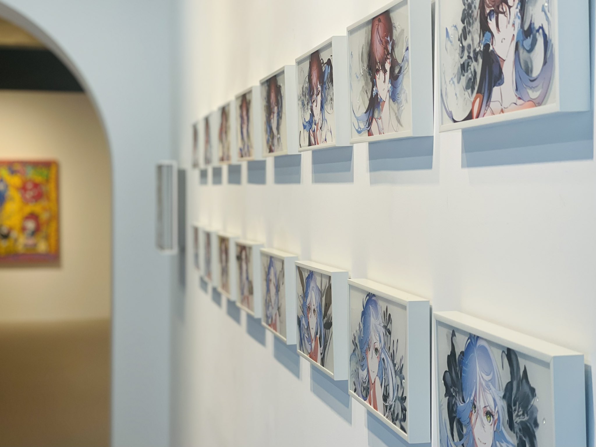 the image shows a hallway in an art gallery with animestyle artwork displayed on the wall. the artwork features female characters with blue hair and blue and white clothing. the hallway is welllit with artificial lighting.