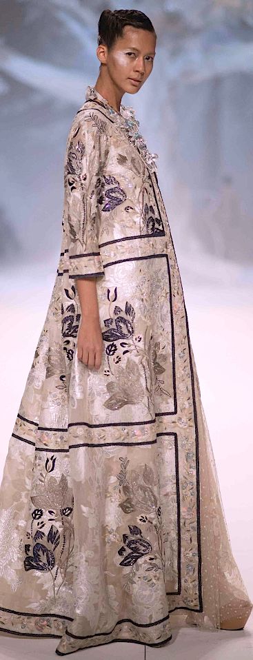 full shot of a fashion model showcasing a long, elegant beige gown. the dress features intricate embroidery with floral and other artistic designs in shades of purple and beige, creating a delicate and luxurious look. the model is asian, with her hair styled up and makeup done subtly. the background is out of focus, suggesting a runway or photoshoot setting. the overall style exudes high fashion and sophistication.