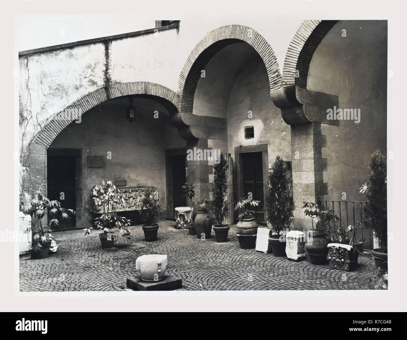 a black and white photograph shows a courtyard in a historical building, likely in italy. the courtyard is paved with small stones arranged in a circular pattern. the building features several arches made of stone, with potted plants and other greenery arranged throughout the courtyard. the architecture appears old, with a sense of history and grandeur.