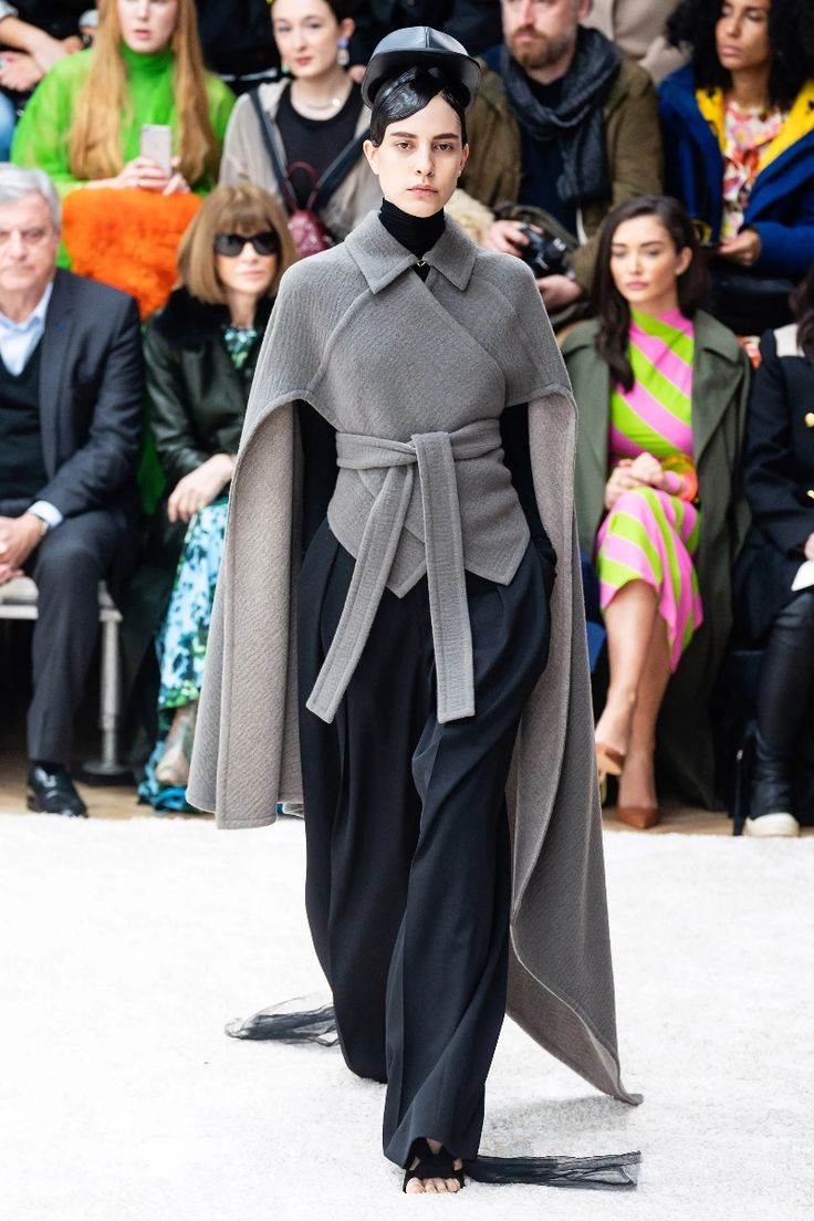a female model walks the runway wearing a gray cape, a matching gray top, and black pants. she has her hair pulled back in a low ponytail, and she is wearing a black hat. her shoes are black sandals with black tassels. the model is looking straight ahead as she walks.