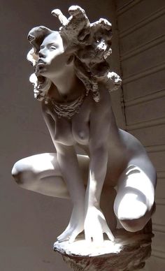 a white marble statue of a woman in a crouching pose with her head turned to the side. she is nude and has a crown of leaves or flowers on her head. the statue is set on a stone base and appears to be a classical piece of art.