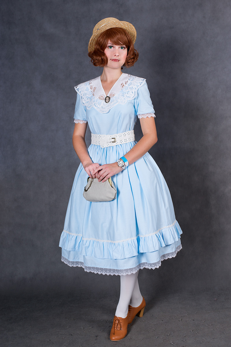 a young woman with shoulderlength auburn hair is modeling a light blue vintagestyle dress.  the dress features a lace collar, a ruffled hemline, and a white belt. she is also wearing a straw hat, brown oxfords, and white socks. she carries a light grey handbag and is posed in a studio setting against a grey backdrop. the overall style is reminiscent of the 1920s or 1930s. the image is a fullbody portrait, and she appears elegant and charming.
