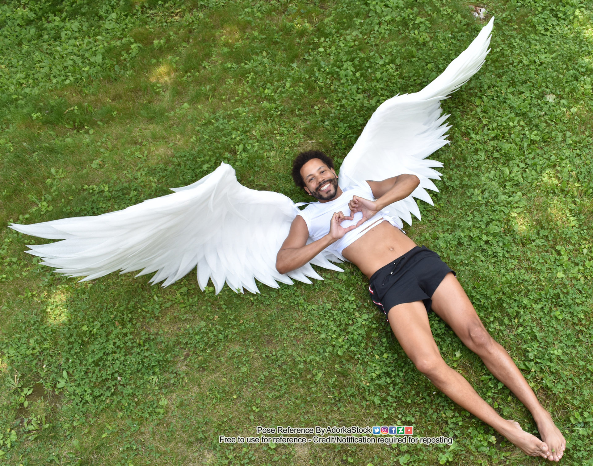 a man with white angel wings is lying on his back in the grass, smiling. he is making a heart shape with his hands. his face is lit up with joy, and he appears to be very happy. the grass is green and lush, and the setting is outdoors. this is a great stock photo for use as a reference for artists or photographers.