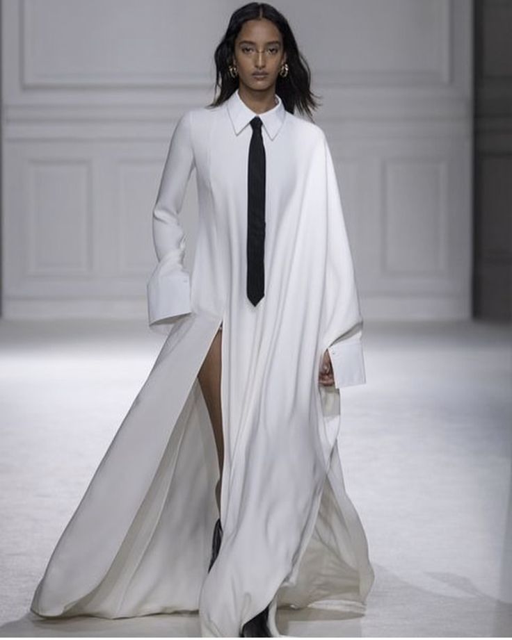 a woman is walking down a runway wearing a white, longsleeved dress with a black tie. the dress has a collar and cuffs similar to a shirt. the dress also features a high slit. the woman is wearing earrings and has dark hair.