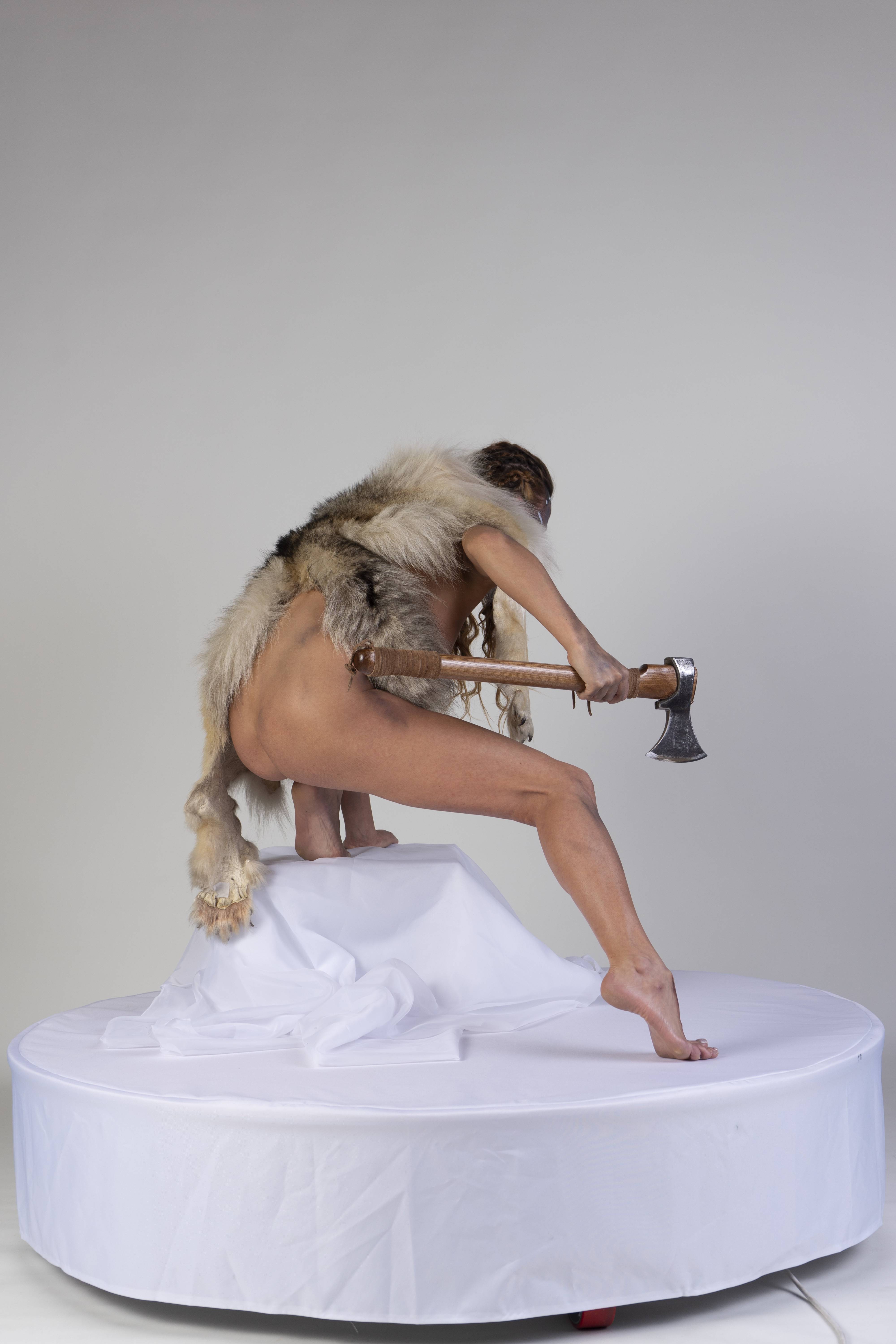 a nude woman wearing fur on her back stands on a white platform and holds an axe behind her back.