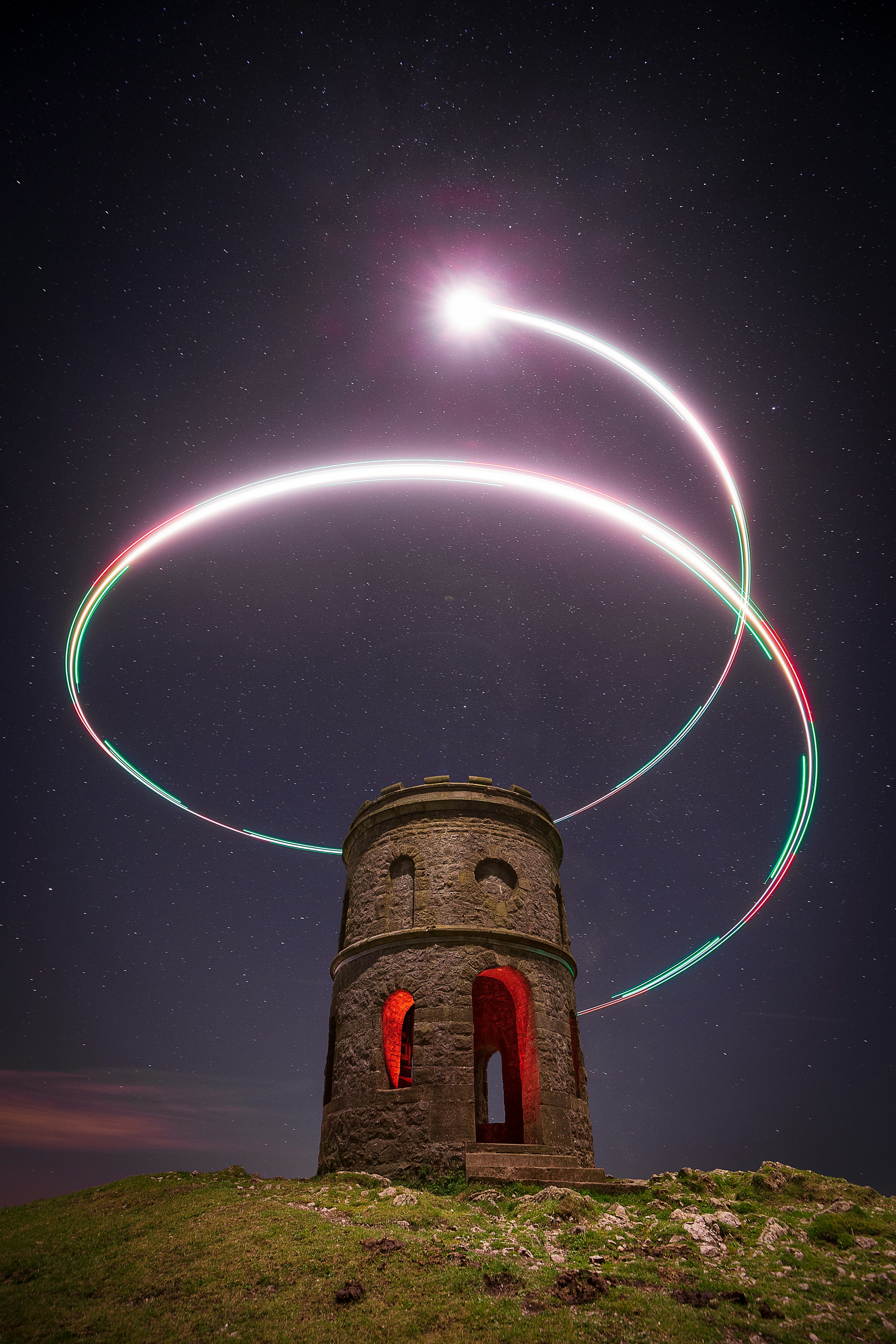 a stone tower stands on a hilltop against a backdrop of a starry night sky. two longexposure light trails, created by a moving light source, encircle the tower, adding a dynamic and otherworldly element to the scene.