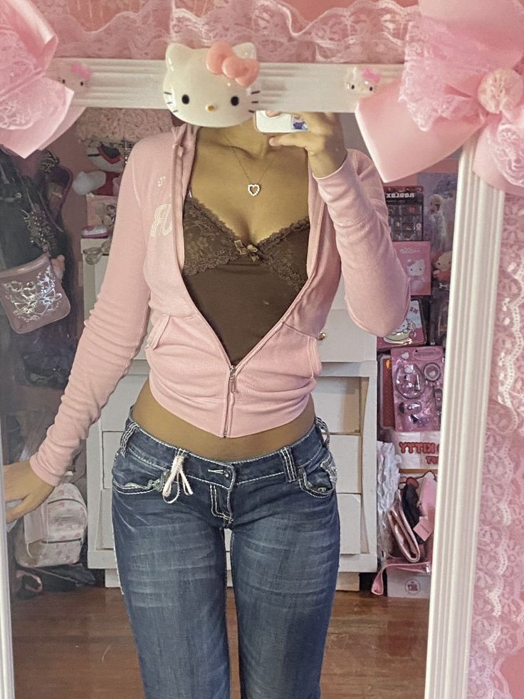 a mirror selfie shows a young woman wearing a light pink juicy couturestyle hoodie with a brown lace bralette underneath. she's also wearing lowrise dark wash jeans. the background is a very pink and girly bedroom with hello kitty decor and lace accents. the overall aesthetic is very y2k and reminiscent of early 2000s fashion trends.