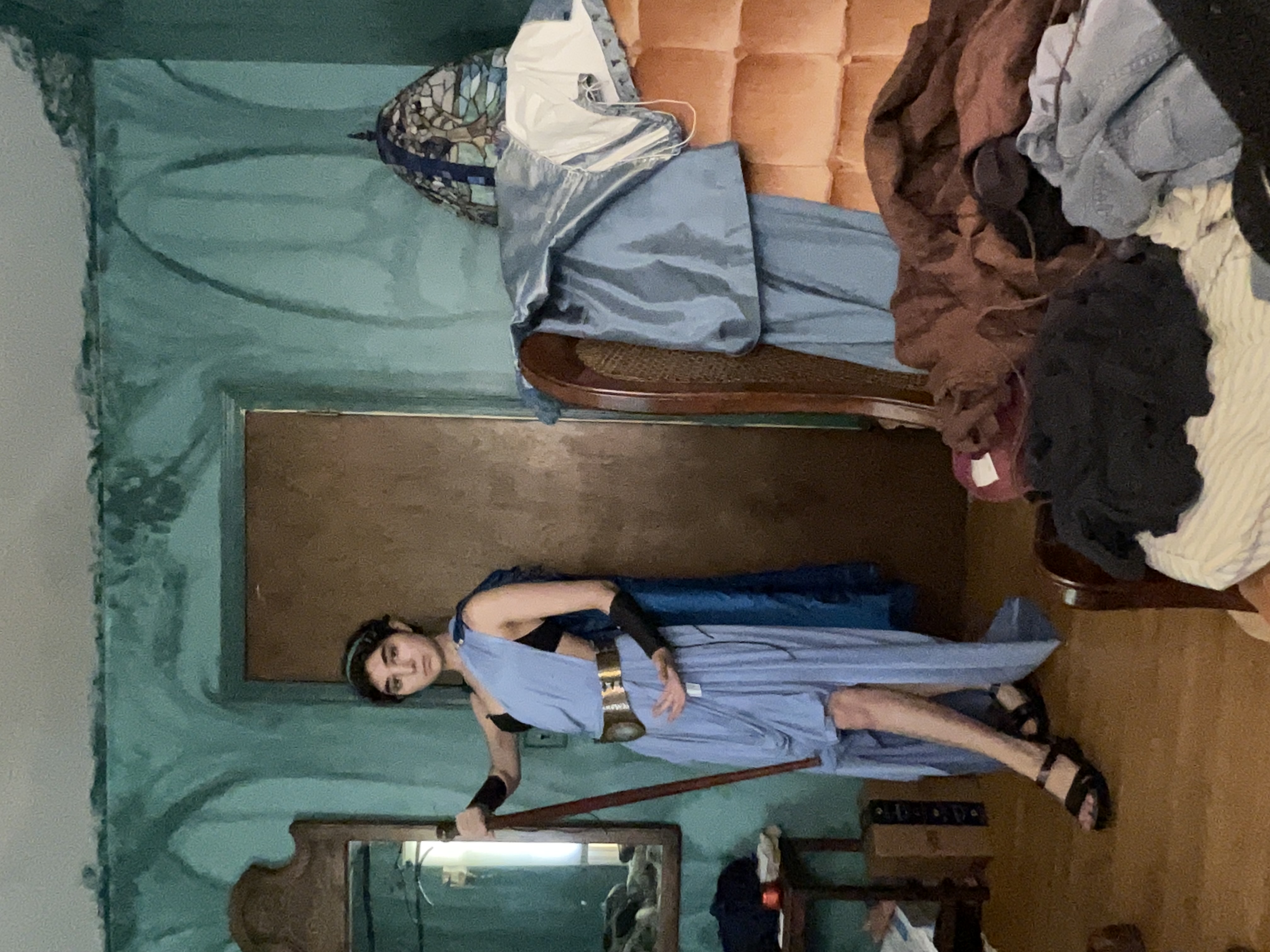 a fulllength indoor shot shows a young woman cosplaying as a greek warrior or goddess. she's wearing a light blue flowing gown with a gold belt, dark sandals, and a dark headband.  she holds a long wooden staff or spear. the setting appears to be a bedroom with teal walls, a vintagestyle chair draped in fabrics, and a messy overall appearance.  a stainedglass lamp is visible in the background.  the woman stands confidently, looking directly at the camera.