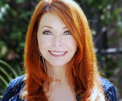a closeup headshot of a smiling woman with long, wavy red hair. she is wearing a blue blouse and has a warm, friendly expression on her face. the background is blurred, likely foliage.