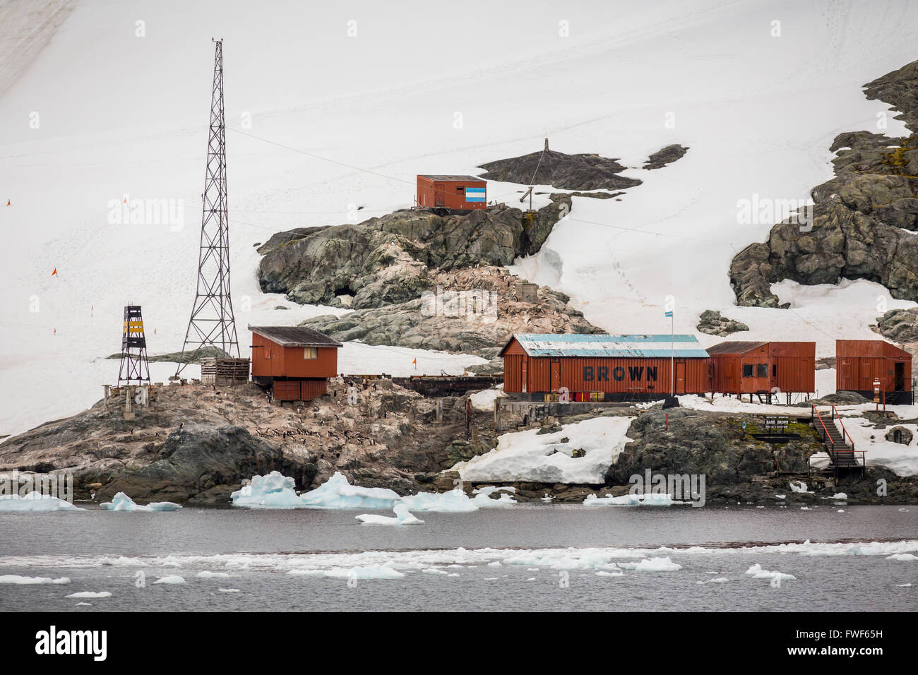 a view of the brown research station in antarctica, the station is surrounded by snowcovered mountains, the station consists of several red wooden buildings, one of the buildings has the words "brown" painted on its side, there is an argentine flag flying on one of the buildings, there is a large tower in the foreground.  the water in the foreground is covered with ice.