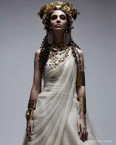 a woman wearing a flowing white gown with intricate gold jewelry stands in a studio, posing for a portrait. she has long curly dark hair adorned with a gold crown and her makeup is sophisticated. the image evokes a sense of classic beauty and elegance, and hints at a  mythological or goddesslike inspiration.