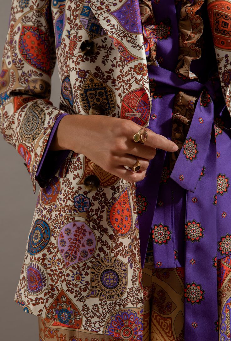 a closeup shot focuses on a woman's torso, showcasing a detailed paisleyprinted silk blazer in cream, orange, purple, and brown hues.  the blazer is doublebreasted and features an intricate floral and geometric pattern. underneath, a vibrant purple silk shirt with a similar paisley design is visible, and a purple sash is tied at the waist. the woman's hands are visible, adorned with gold rings. the overall aesthetic is vintageinspired, with a bohochic feel. the image emphasizes the rich colors and textures of the fabrics.