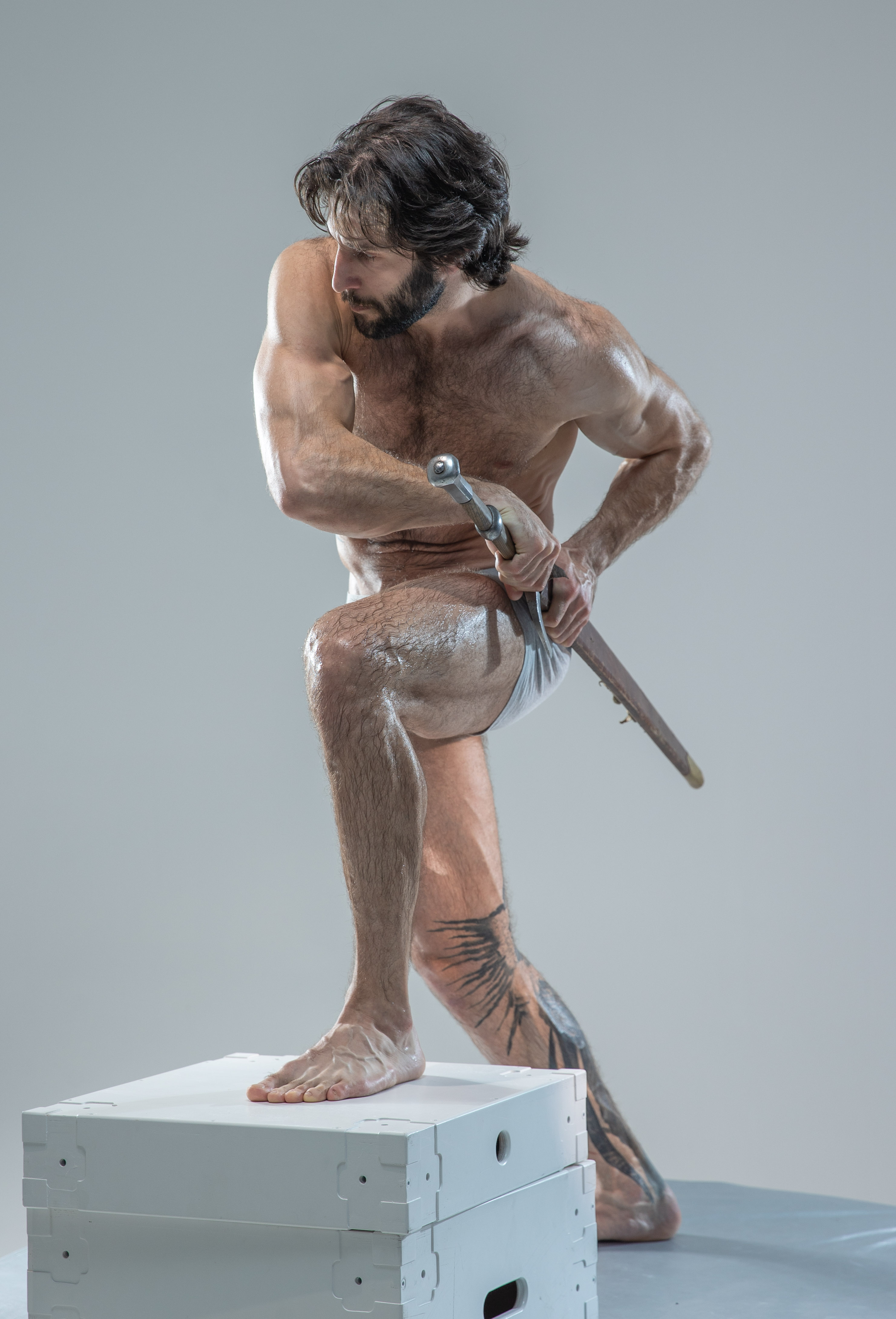 the image showcases a muscular, bearded man in a dynamic pose, reminiscent of a classical sculpture. he stands on a white platform, his body glistening, suggesting oil or sweat. he holds a sword with a determined expression, suggesting action or combat. the background is a neutral gray, highlighting the man's physique and the sword's detail. the lighting is studioquality, casting soft shadows and accentuating his musculature.