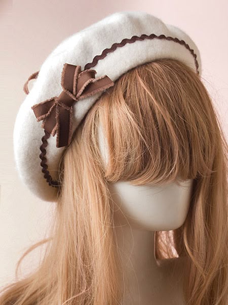 a creamcolored beret with a brown ribbon and scalloped trim sits on a mannequin with a light brown wig.