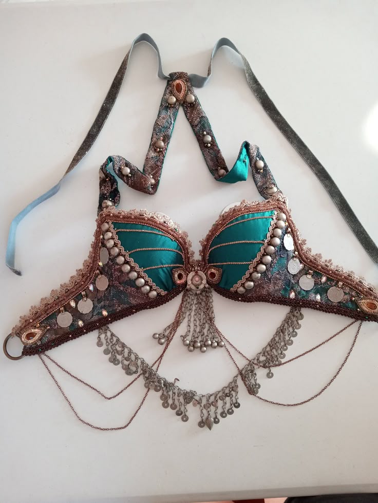 a detailed shot of a belly dance bra on a white surface. the bra is turquoise with intricate beadwork and coin embellishments. a matching neck strap with similar embellishments is attached. the bra has chains and coin dangles hanging from the bottom.