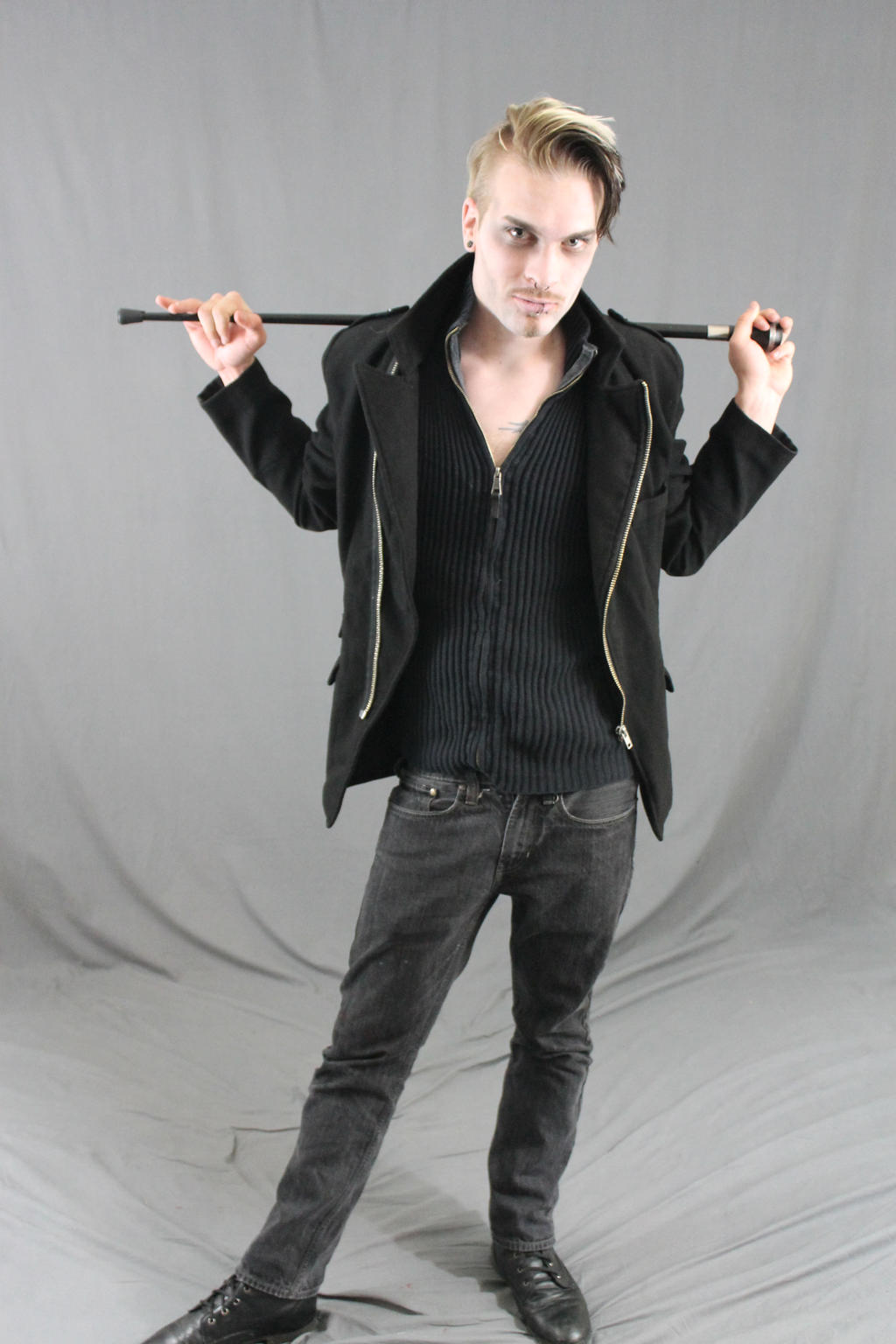 a full shot of a young man with blond hair, pale skin, and dark makeup posing against a gray backdrop. he wears a black coat, a black sweater, dark jeans, and black shoes. he holds a black cane across his shoulders, and his expression is serious yet confident. the overall style is elegant and stylish, with a touch of gothic or alternative flair.