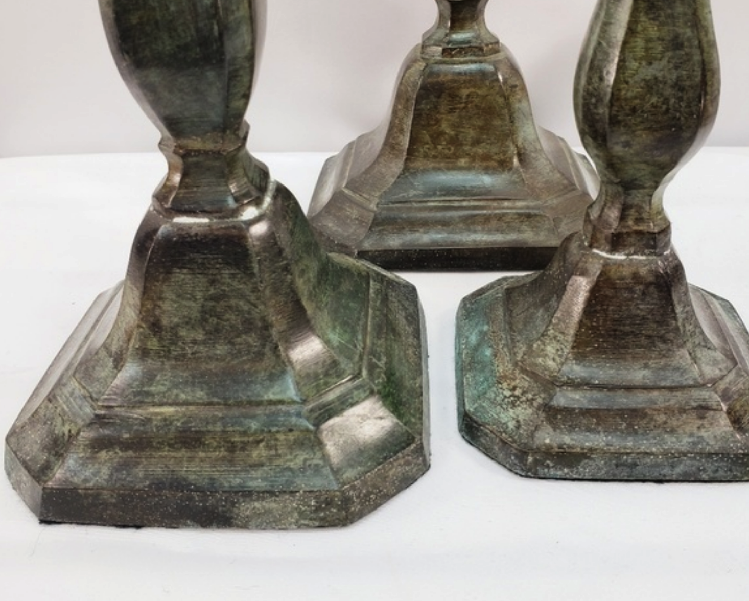 three antique candlestick holders made of metal. the candle holders are a weathered green/grey/silver color and have a rustic look. they are all similar in design but slightly different in size. the candle holders are sitting on a white surface.
