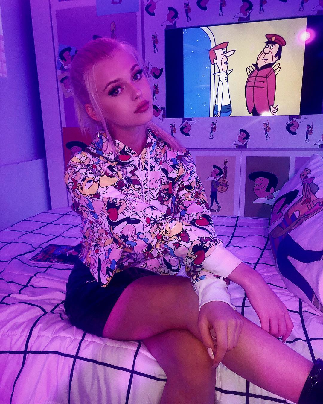 a young woman with blonde hair sits crosslegged on a bed in a room illuminated with purple lighting. she is wearing a hoodie with a cartoon character pattern and dark shorts. behind her is a tv screen showing a cartoon. the bed has a grid pattern bedspread. the walls are decorated with cartoonthemed posters and wallpaper.