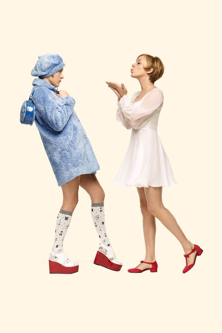 two women are standing sidebyside. the woman on the left is wearing a blue dress, white kneehigh socks with black patterns and red wedge platform shoes. she is holding a blue bag. the woman on the right is wearing a white dress and red sandals. she is smiling and sending a kiss to the camera. they are both posed in a fashion studio with white background.