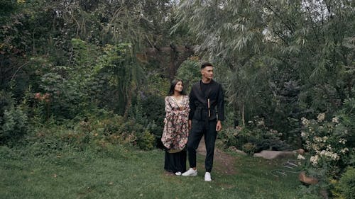 a man and woman stand together in a lush green park. the woman is wearing a floral dress and the man is wearing black clothing. they are looking away from the camera.  the scene is romantic and peaceful. the couple appears to be of asian descent.