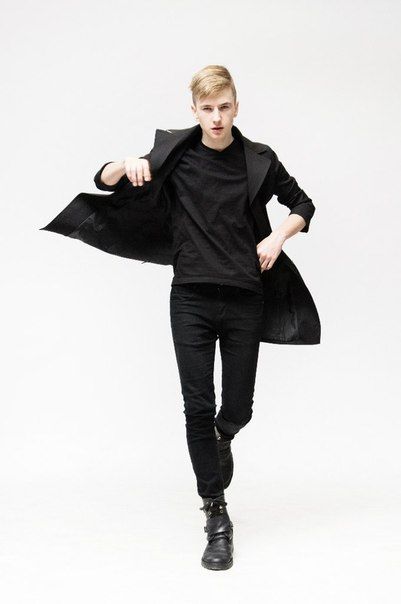 a young man wearing black clothing stands in front of a white background. he has on a black blazer, black jeans, and black boots. he's holding his blazer open and is looking towards the camera.