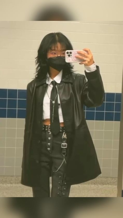 a young girl wearing a black leather jacket, white shirt, black tie, and black pants. she has a silver chain hanging from her belt and is posing for a selfie in a bathroom mirror. her look is cool and edgy.