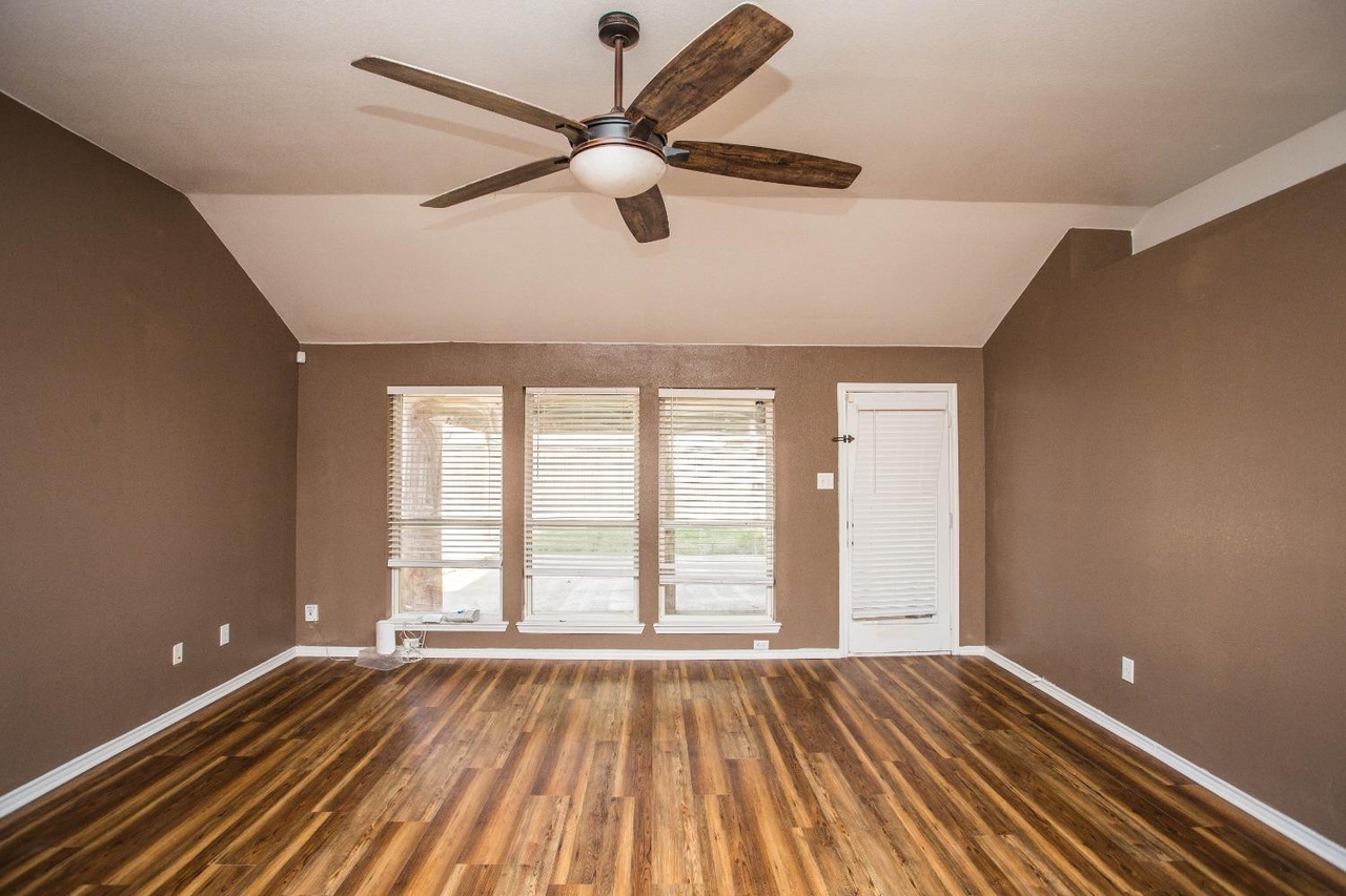 an empty room with hardwood floors, brown walls, a ceiling fan, windows with blinds, and a door.