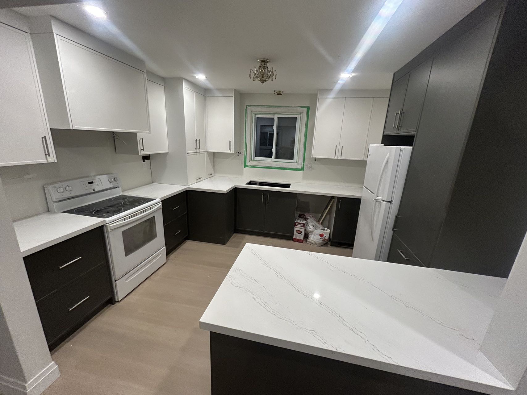 the image shows a newly renovated kitchen. the kitchen features twotoned cabinets, with white upper cabinets and dark lower cabinets.  the countertops are a white quartz with gray veining.  the appliances are white, and there's a kitchen island with the same quartz countertop. the flooring is lightcolored hardwood. recessed lighting illuminates the space, and a chandelier hangs above. the kitchen is clean and modern. the image suggests the renovation is near completion, with a few items still on the floor.