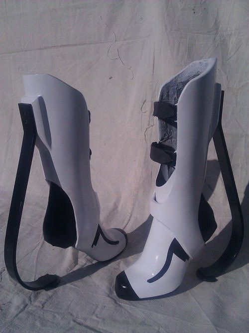 a pair of white and black futuristic boots that appear to be a prop or cosplay item. they have a distinct design with high shins, open toes, and black accents. the boots have buckles and straps and are standing on a white surface, likely indoors under bright, natural lighting.