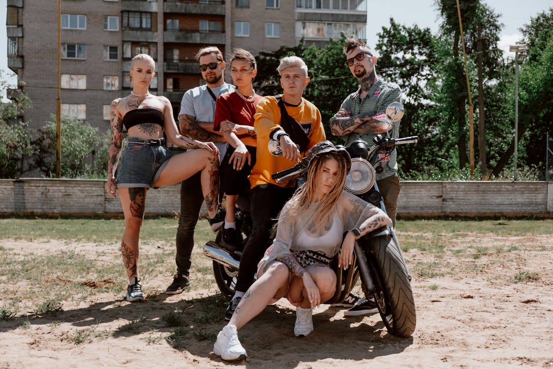 the image shows a group of six young adults,  three men and three women,  all with visible tattoos,  standing and sitting around a vintagestyle motorcycle in an urban setting. the background features a city building and trees. the group is dressed in a casual, alternative style with a mix of  stylish clothing.  they appear relaxed and confident in their pose. the overall mood is one of friendship and a rebellious, cool attitude. the photograph has a slightly desaturated color palette giving it a vintage feel.