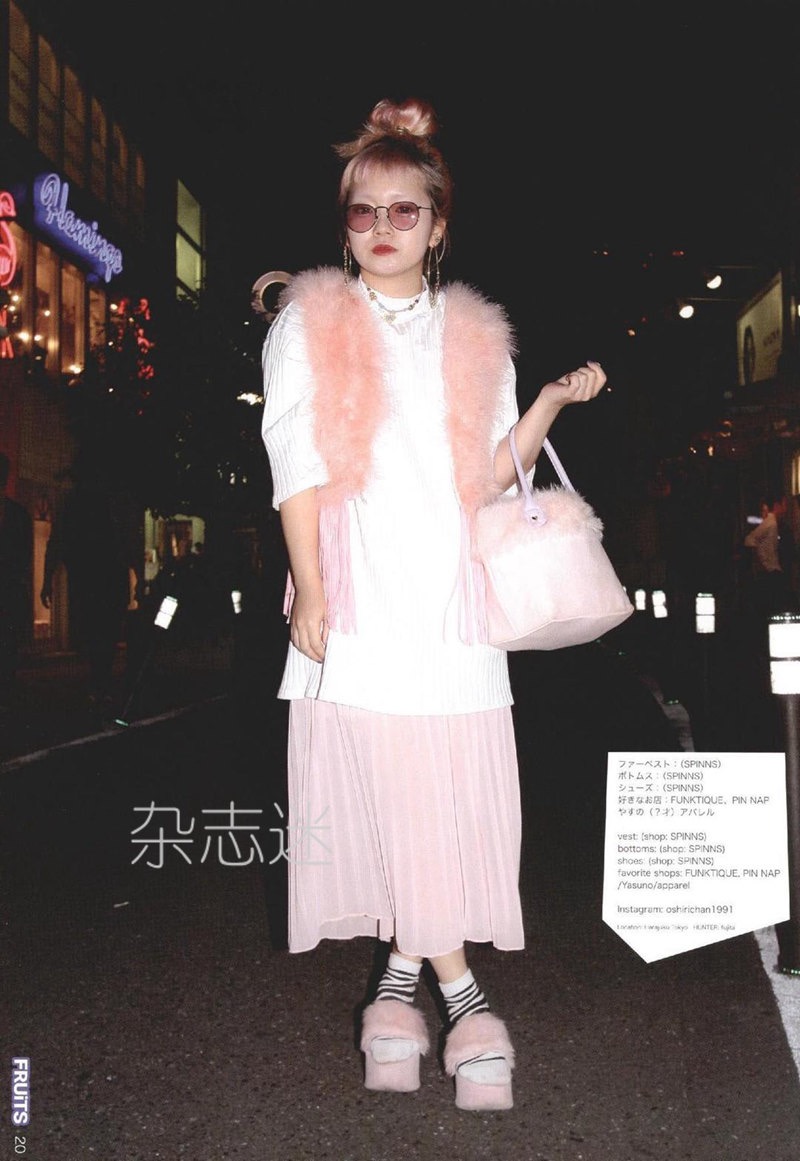 a young woman is posing on a city street at night. she has pink hair styled in a bun and is wearing a pink and white outfit with fluffy pink accessories. the photo has a street style, fashionforward vibe. the background suggests an urban setting, possibly in japan.