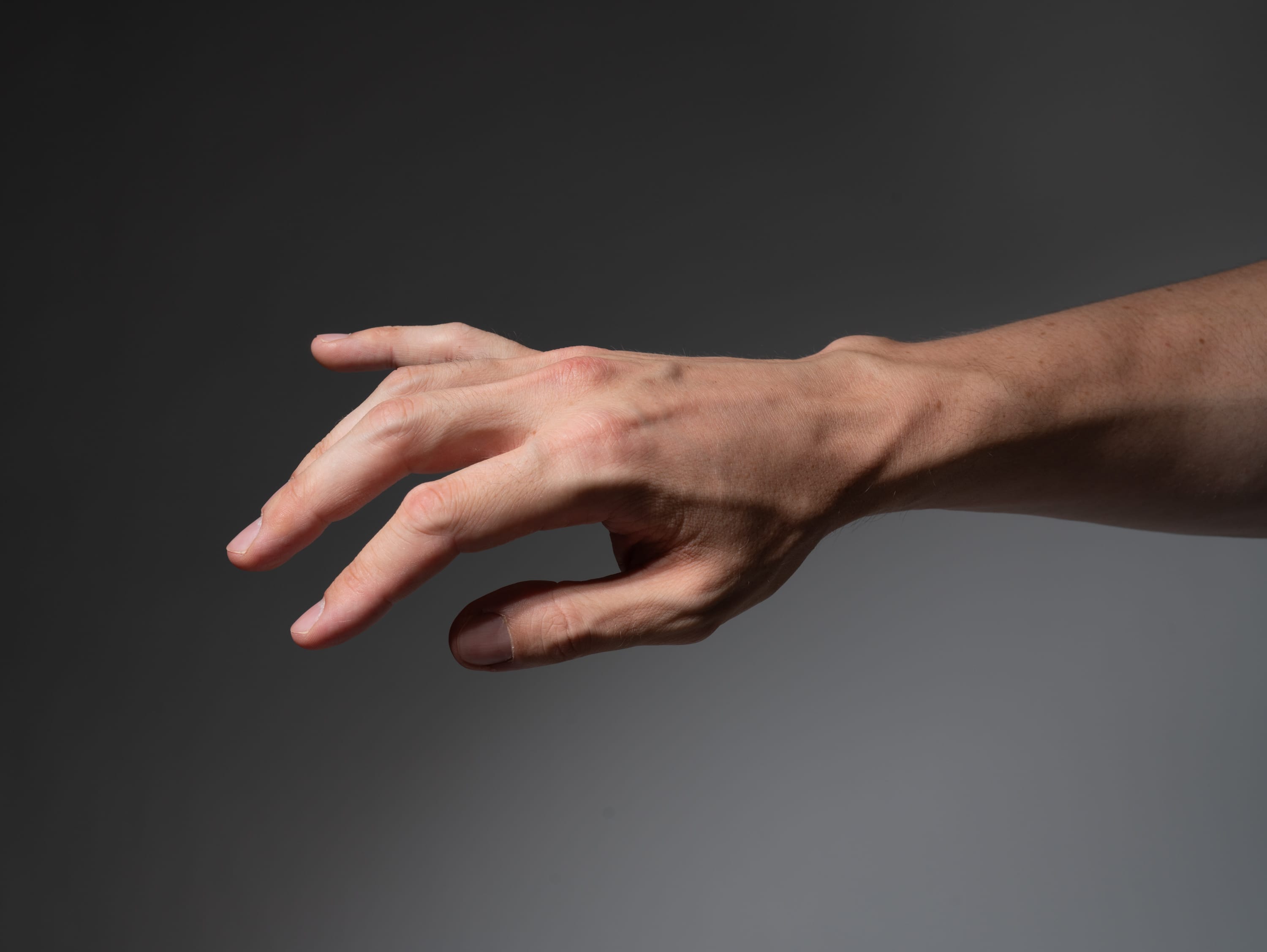 a man's right hand is reaching out against a gray background, illuminated with studio lighting. the image showcases the hand's details, including veins and skin texture, in a side view perspective.