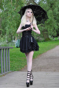 a young, slender woman with long blonde hair is pictured standing on a wooden walkway in a park. she's wearing a black gothic lolitastyle dress with lace accents and black platform laceup boots.  she carries a black lace parasol and a small green bag. the setting is outdoors, with lush green trees in the background. the overall style is gothic and alternative fashion.