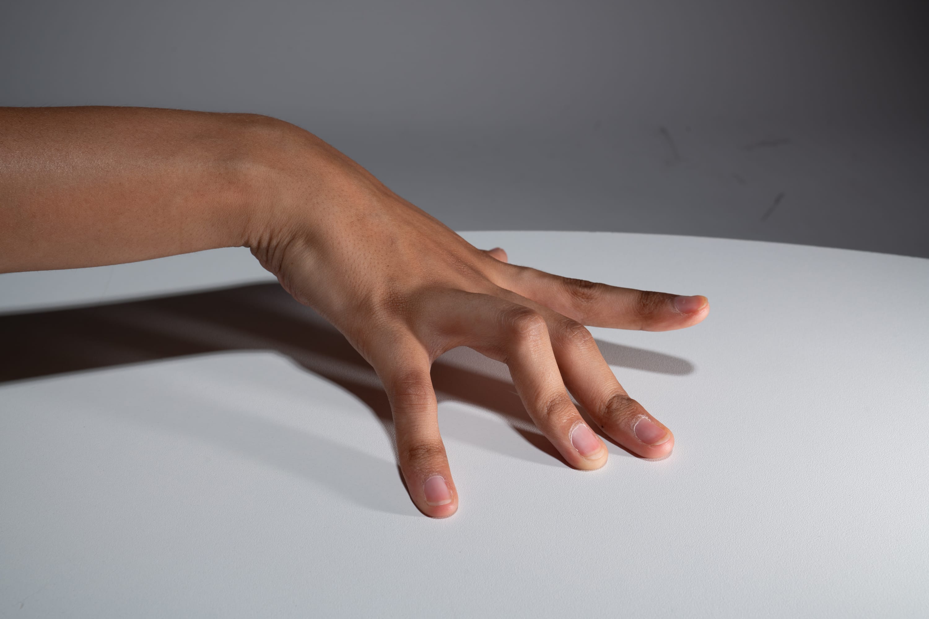 a hand with its fingers spread out rests on a white table. the hand is positioned horizontally with the fingers pointing towards the right side of the image. the lighting is soft and diffused, creating subtle shadows on the table.