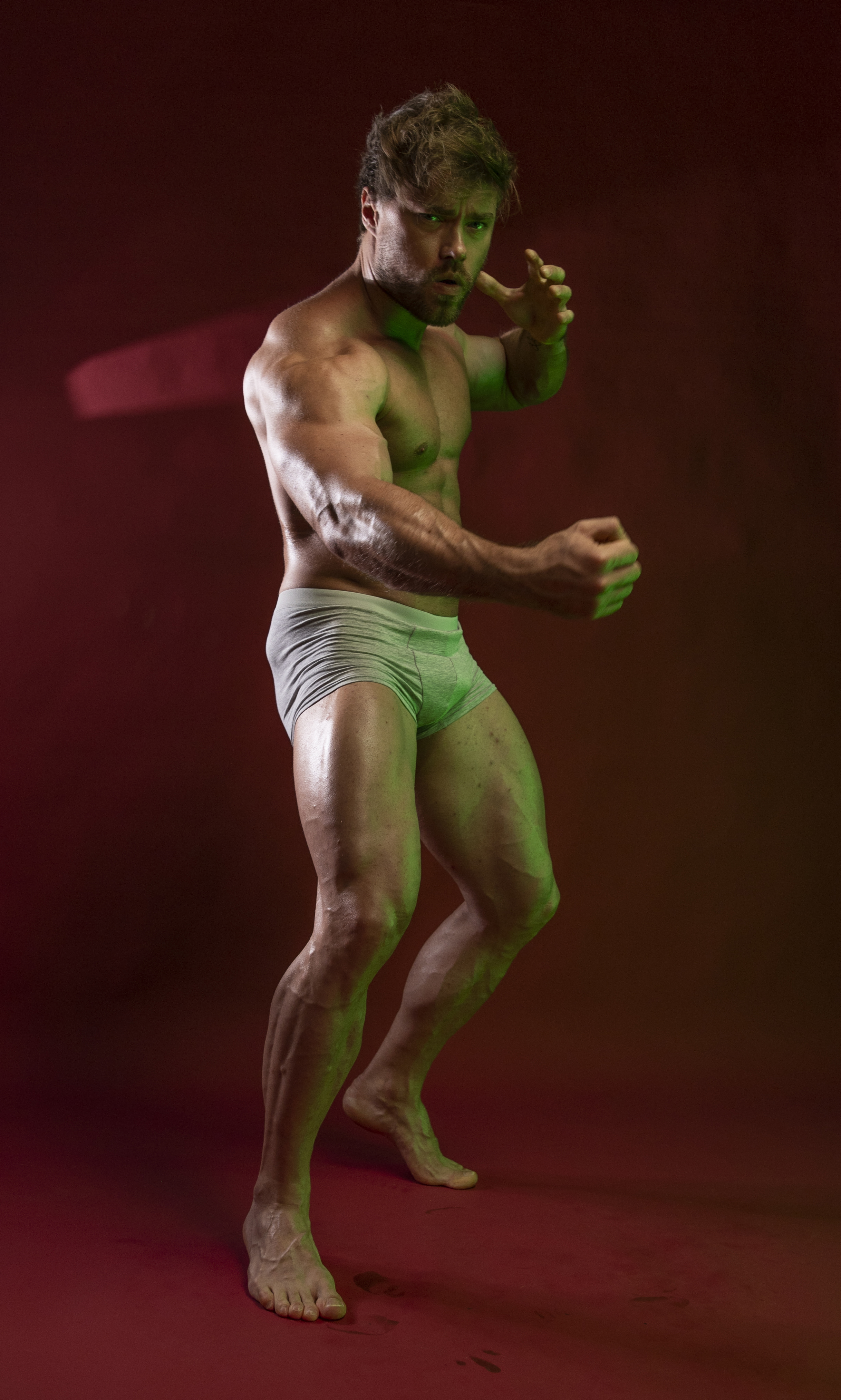 a muscular man is standing shirtless in light gray underwear with bare feet, set against a dark red background. the man has a strong physique, showing prominent biceps, calf muscles, and visible veins. he is illuminated with green lighting, creating a dramatic effect. he appears to be in a fighting stance with his fists clenched and raised. the image is a studio shot taken indoors.