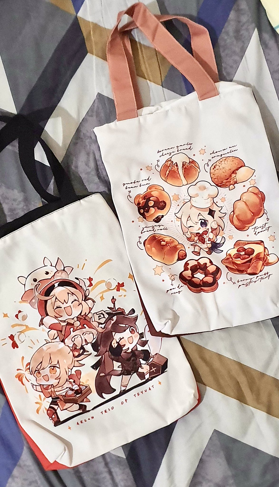the image shows two tote bags lying on a patterned surface. the tote bag on top has an image of various food items and a character from the anime "genshin impact." the tote bag on the bottom has an image of three characters from the anime, "genshin impact."