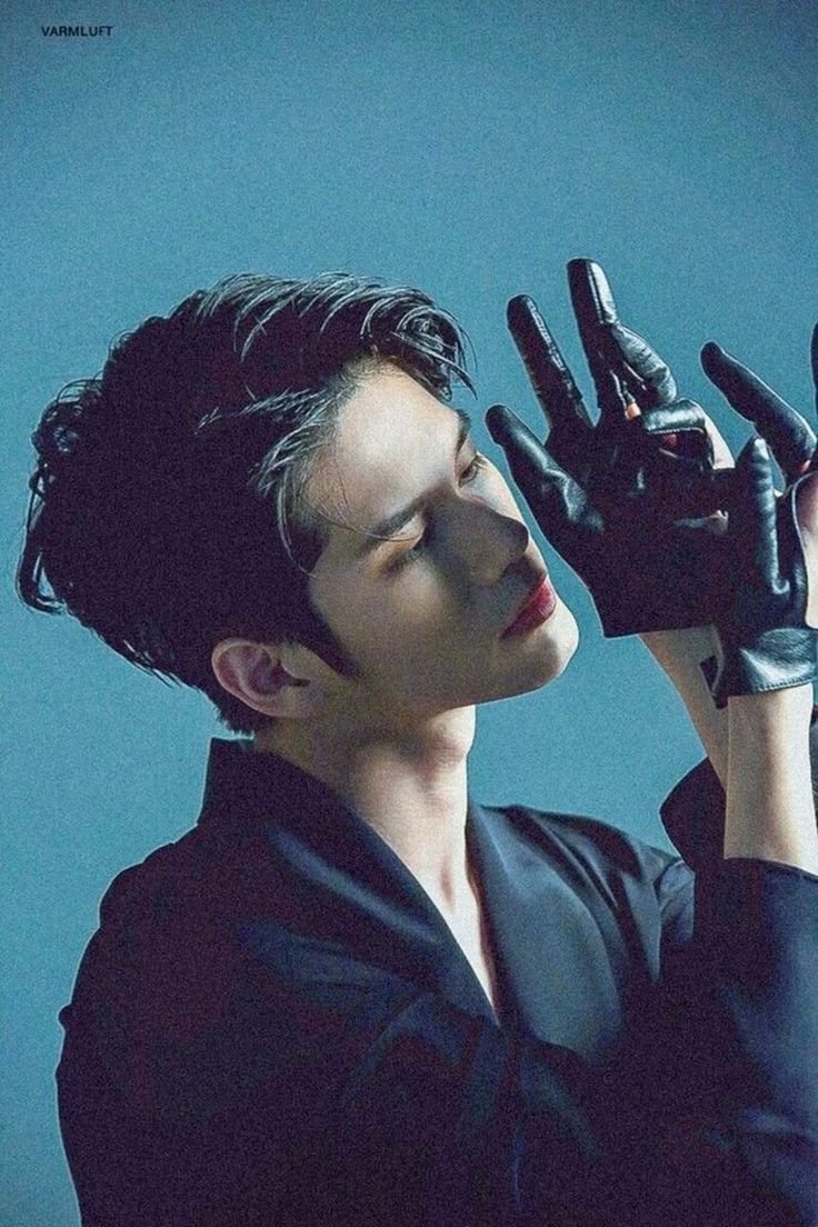 the image features a closeup portrait of a young man with dark hair styled impeccably. he is wearing a black collared shirt and sleek black leather gloves, which he holds up partially obscuring his face. his eyes are closed, head tilted slightly back, and lips slightly parted, creating a sense of drama and allure. the background is a muted teal, providing a cool contrast to the subject's dark attire and intense expression. the soft lighting further enhances the overall mood of sophisticated elegance.