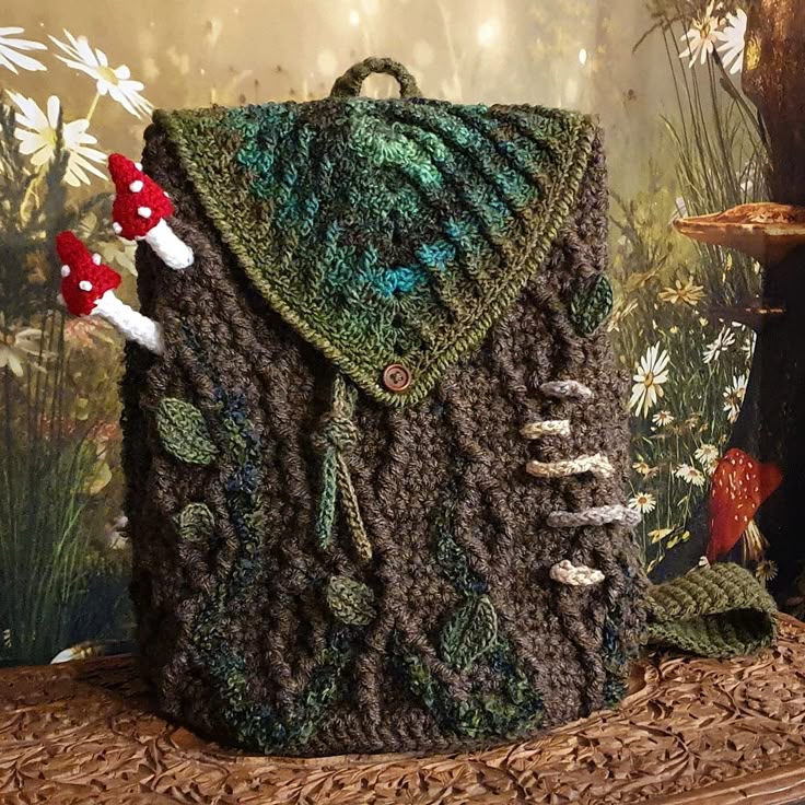a handmade, naturethemed crochet backpack sits on a wooden surface with a backdrop of flowers and foliage. the backpack is predominantly brown and green, resembling tree bark or moss. it features knitted details such as leaves and mushrooms. two red and white mushrooms are attached to the side. the backpack has a flap secured with a button and a knitted drawstring closure. a small knitted handle is on top. the overall design evokes a forest or woodland aesthetic.