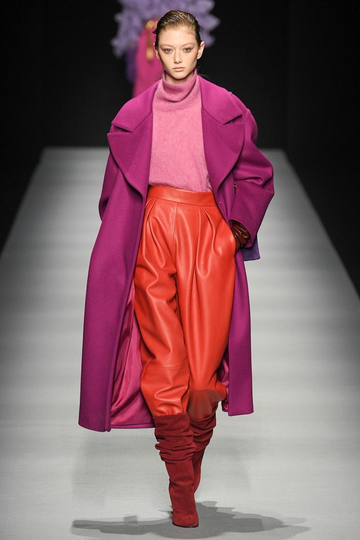 a female fashion model walks down a runway during a fashion show, showcasing a pink and red outfit. she is wearing a long purple coat with a pink turtleneck sweater and bright red leather pants. she also has on red boots that hit above her knees. her hair is styled in a simple straight style and she is wearing minimal makeup. the background of the image shows the runway and a black backdrop. the image captures the model's outfit and confident walk down the runway, showcasing the latest in fall/winter fashion trends.