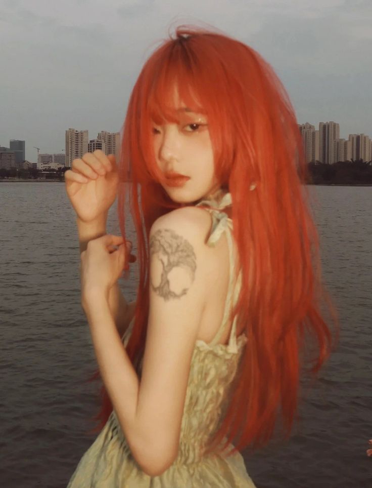 a young woman with long, bright red hair is standing with her back to the camera, looking over her left shoulder. she is wearing a pale green dress and has a tree tattoo on her left upper arm. the background is a cityscape with water and buildings in the distance. the photo is lit with warm, natural light, likely from the setting sun.